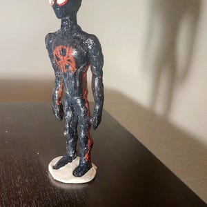 May include: A handmade figurine of a superhero, featuring a black and red color scheme. The figure stands on a white base, with a red spider emblem on the chest. The eyes are white with red outlines.