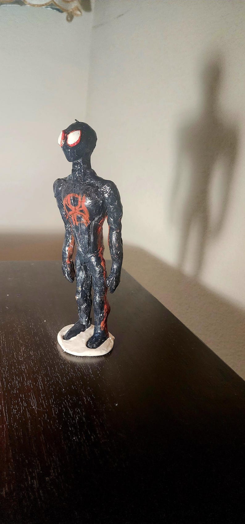 May include: A handmade figurine of a superhero, featuring a black and red color scheme. The figure stands on a white base, with a red spider emblem on the chest. The eyes are white with red outlines.