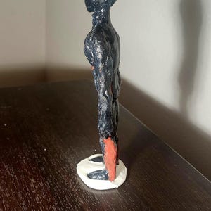 May include: A handmade sculpture of a humanoid figure. The figure is primarily black with red accents on the feet and head. It stands on a white base and has a textured surface.