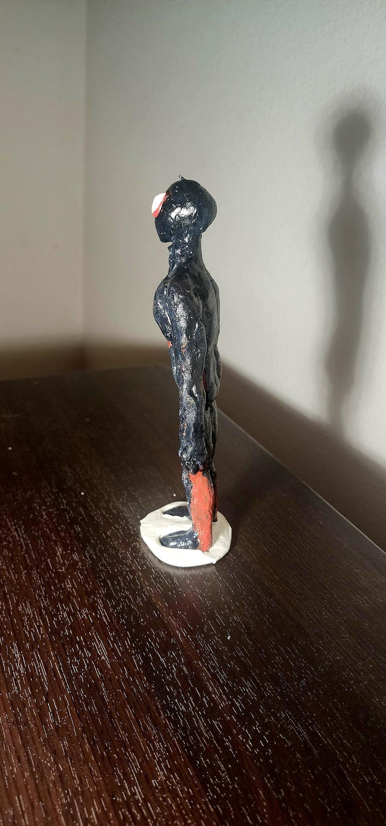 May include: A handmade sculpture of a humanoid figure. The figure is primarily black with red accents on the feet and head. It stands on a white base and has a textured surface.