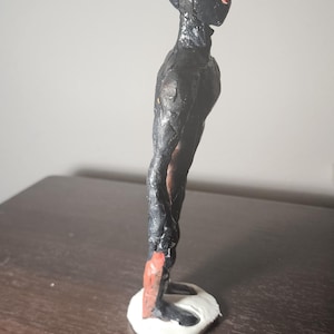 May include: A handmade sculpture of a humanoid figure with a black body and head, and a red accent on the face. The figure stands on a white base. The sculpture is on a dark brown surface.