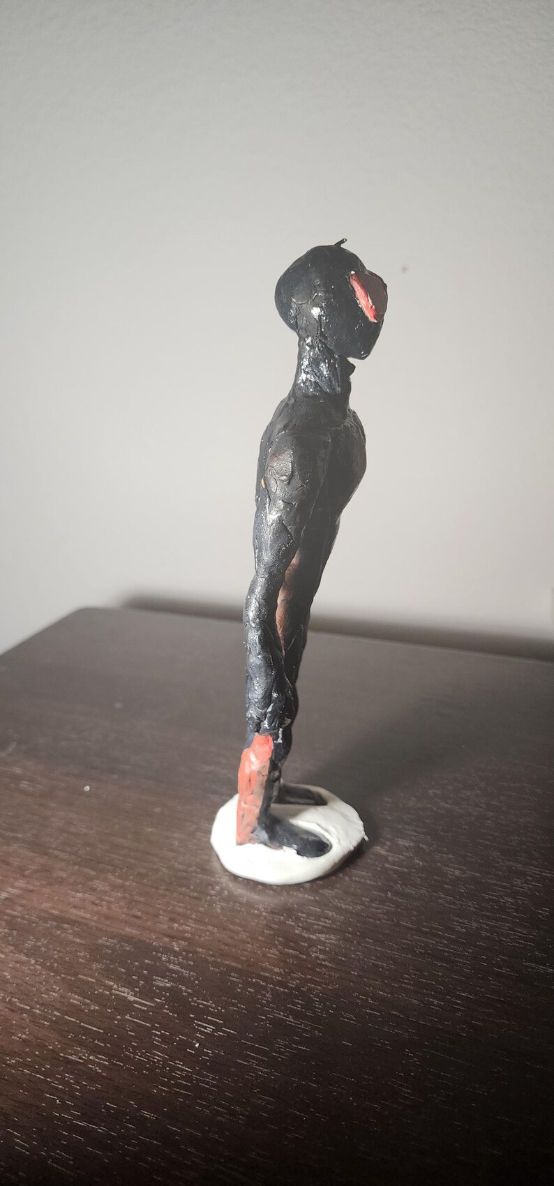May include: A handmade sculpture of a humanoid figure with a black body and head, and a red accent on the face. The figure stands on a white base. The sculpture is on a dark brown surface.