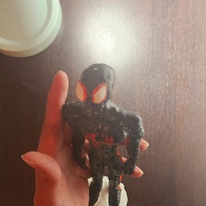 May include: A handmade black and red figurine of a superhero, with large red eye shapes. The figure is standing on a white base. The figurine is held in a hand against a dark brown background.