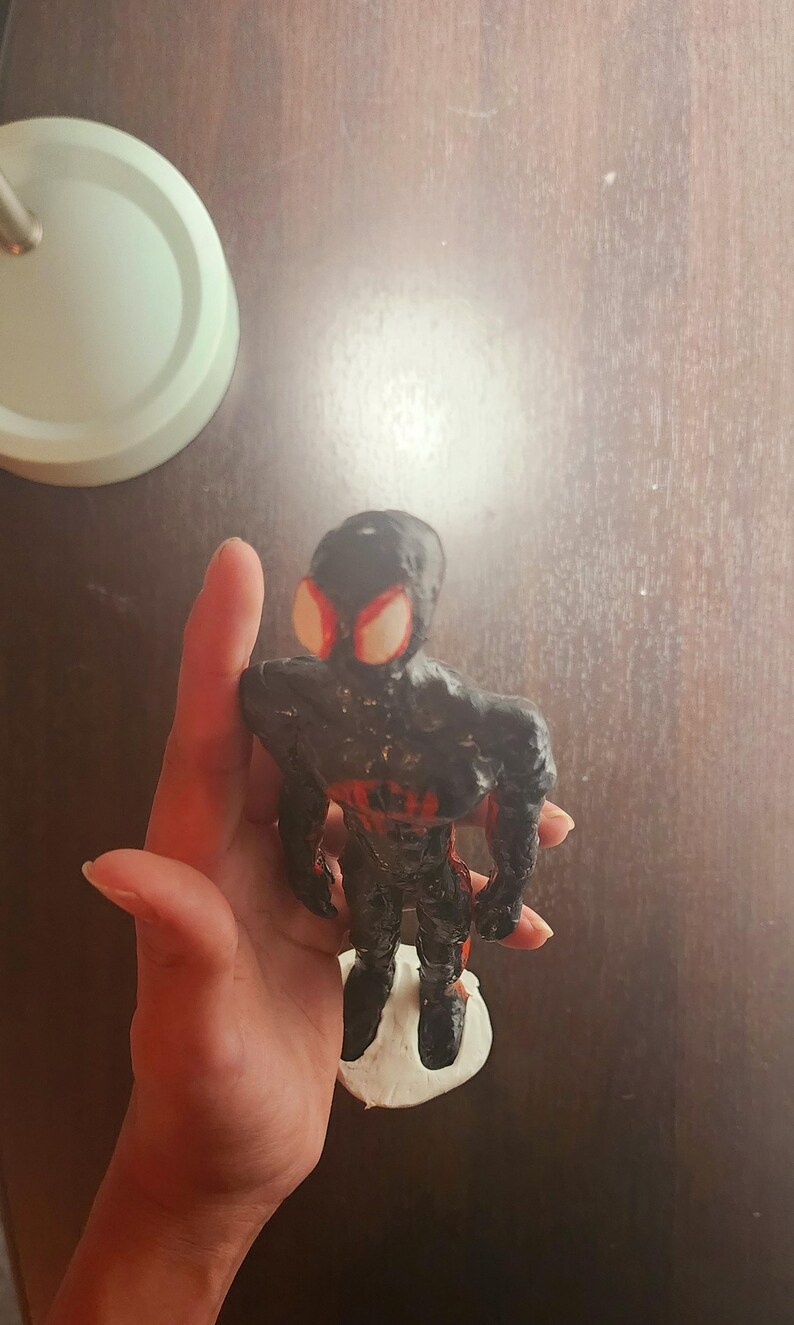May include: A handmade black and red figurine of a superhero, with large red eye shapes. The figure is standing on a white base. The figurine is held in a hand against a dark brown background.
