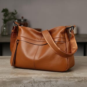 Peut inclure: Un sac à bandoulière en cuir marron avec une surface texturée. Le sac a une longue bandoulière réglable et une poignée plus courte. Le sac est posé sur une surface grise.