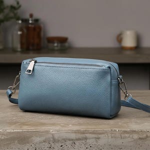 May include: A light blue rectangular crossbody bag with a silver zip and strap hardware. The bag has a textured leather appearance and a long, thin strap. The bag is on a concrete surface.