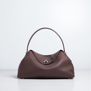 Puede incluir: Bolso de mano de cuero marr&oacute;n oscuro con asa curva. El bolso tiene forma redondeada con cierre de solapa y cierre plateado. El cuero tiene un aspecto texturizado.