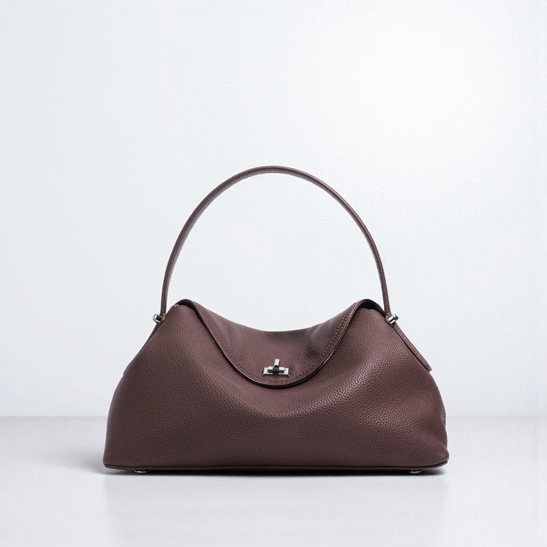 Puede incluir: Bolso de mano de cuero marr&oacute;n oscuro con asa curva. El bolso tiene forma redondeada con cierre de solapa y cierre plateado. El cuero tiene un aspecto texturizado.