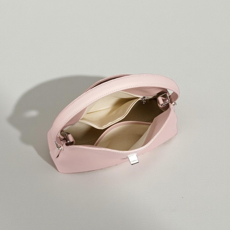 Puede incluir: Un bolso de mano rosa claro con un asa curva y un cierre plateado. El interior presenta m&uacute;ltiples compartimentos con un forro beige claro. El bolso est&aacute; hecho de un material liso similar al cuero.
