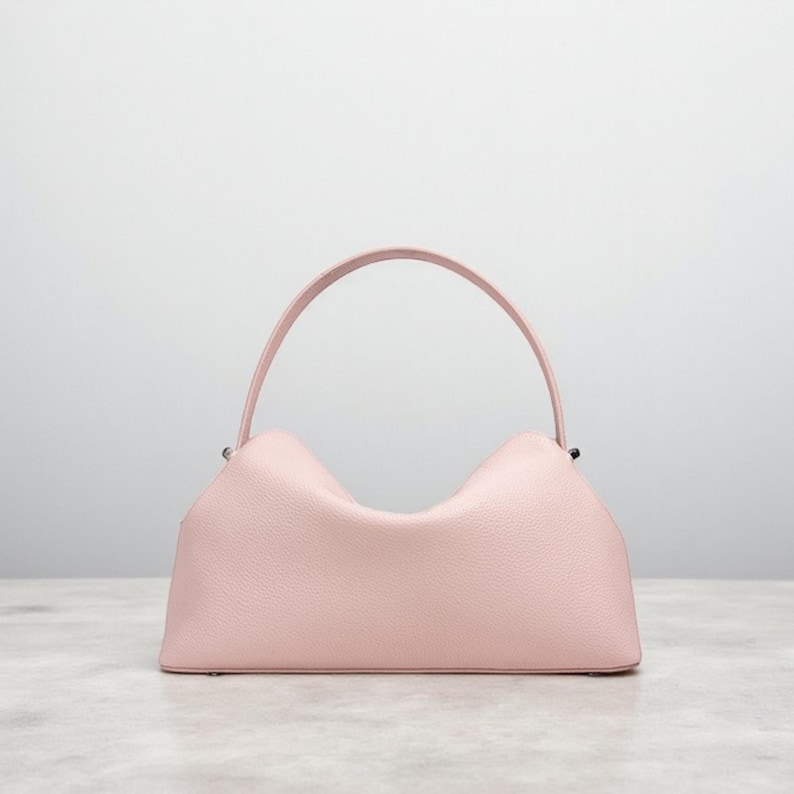 Puede incluir: Un bolso de mano rosa claro con un asa curva. El bolso tiene un cuerpo rectangular con un dise&ntilde;o superior &uacute;nico en forma de ola. El asa es de color rosa claro a juego, y el bolso parece estar hecho de un material texturizado.