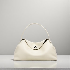 Puede incluir: Bolso de mano de cuero color crema con una parte superior curvada y un solo asa. El bolso tiene una forma estructurada con una textura sutil y un peque&ntilde;o cierre plateado. El asa se arquea elegantemente sobre el cuerpo principal del bolso.