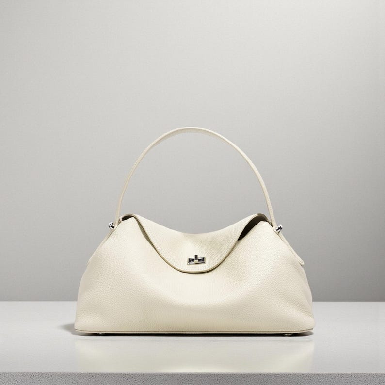 Puede incluir: Bolso de mano de cuero color crema con una parte superior curvada y un solo asa. El bolso tiene una forma estructurada con una textura sutil y un peque&ntilde;o cierre plateado. El asa se arquea elegantemente sobre el cuerpo principal del bolso.
