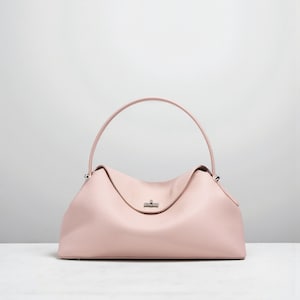 Puede incluir: Un bolso de mano de cuero rosa claro con un asa curva. El bolso tiene una forma estructurada con un asa superior y un peque&ntilde;o cierre plateado. El cuero tiene una apariencia texturizada y el bolso est&aacute; sobre una superficie blanca.
