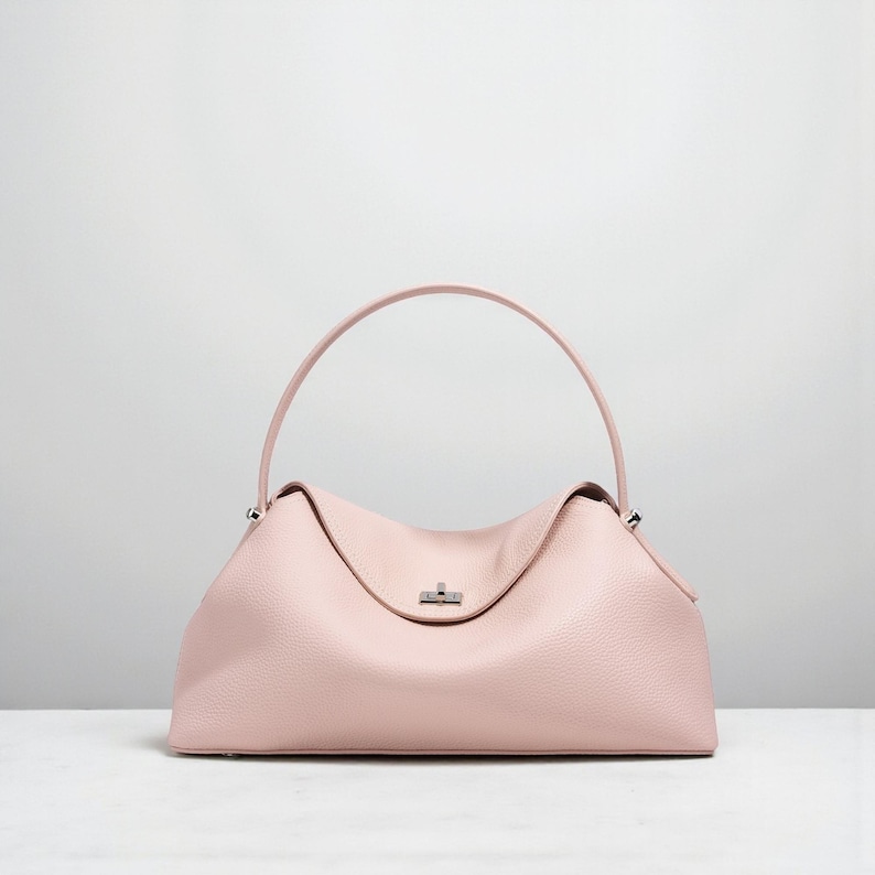 Puede incluir: Un bolso de mano de cuero rosa claro con un asa curva. El bolso tiene una forma estructurada con un asa superior y un peque&ntilde;o cierre plateado. El cuero tiene una apariencia texturizada y el bolso est&aacute; sobre una superficie blanca.