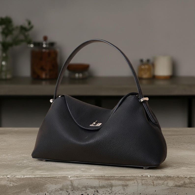 Puede incluir: Un bolso de mano de cuero negro con un asa curva. El bolso tiene una forma triangular &uacute;nica con un cierre de solapa y herrajes plateados. El cuero texturizado le da al bolso un aspecto cl&aacute;sico. El bolso est&aacute; sobre una superficie de hormig&oacute;n.