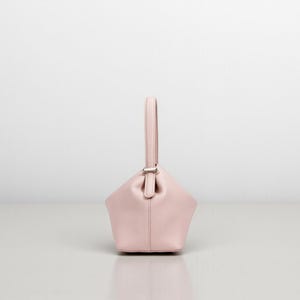 Puede incluir: Un bolso de mano rosa claro con una forma geom&eacute;trica &uacute;nica y un asa individual. El bolso presenta un cierre plateado y est&aacute; hecho de un material liso. Se encuentra sobre un fondo blanco liso, resaltando su dise&ntilde;o.