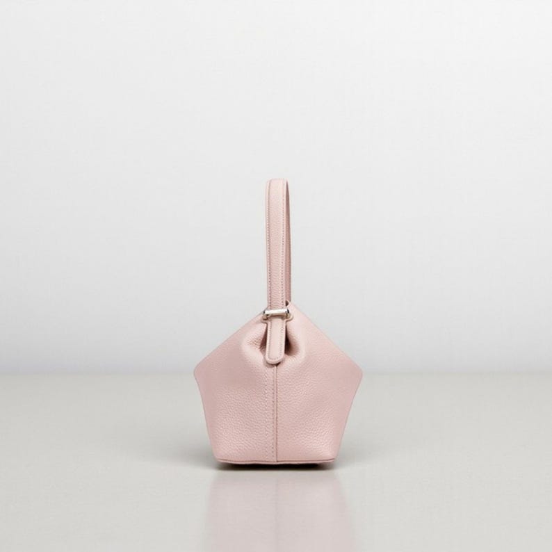Puede incluir: Un bolso de mano rosa claro con una forma geom&eacute;trica &uacute;nica y un asa individual. El bolso presenta un cierre plateado y est&aacute; hecho de un material liso. Se encuentra sobre un fondo blanco liso, resaltando su dise&ntilde;o.