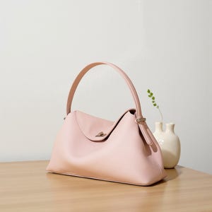 Puede incluir: Un bolso de mano rosa claro con un asa curva y una superficie texturizada. El bolso tiene un cierre de solapa y un cierre plateado. Un peque&ntilde;o jarr&oacute;n blanco con una ramita de vegetaci&oacute;n est&aacute; al lado del bolso sobre una superficie de madera.