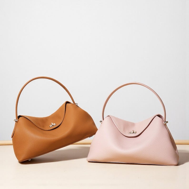 Puede incluir: Dos bolsos de cuero. Uno es de color marr&oacute;n claro, el otro es rosa. Ambos tienen un asa superior curvada y un cierre plateado. Los bolsos tienen forma trapezoidal con la parte superior curvada y la base plana.