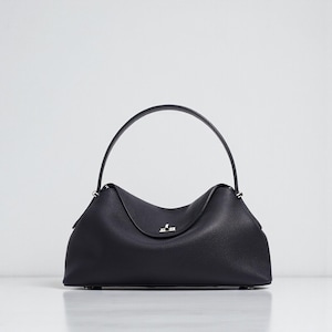 Puede incluir: Un bolso de mano de cuero negro con un asa superior curvada. El bolso tiene una forma estructurada con una superficie sutilmente texturizada. Un cierre plateado est&aacute; centrado en la parte delantera.