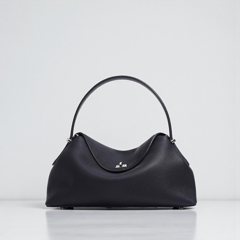 Puede incluir: Un bolso de mano de cuero negro con un asa superior curvada. El bolso tiene una forma estructurada con una superficie sutilmente texturizada. Un cierre plateado est&aacute; centrado en la parte delantera.