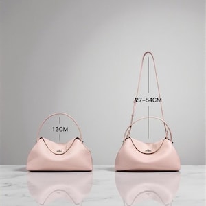 Puede incluir: Dos bolsos de mano de cuero rosa claro. Uno tiene un asa superior y mide 13 cm de ancho. El otro tiene una bandolera y mide 27-54 cm de alto. Ambos bolsos tienen forma curva y un peque&ntilde;o cierre met&aacute;lico.