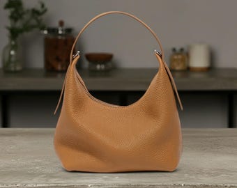 Puede incluir: Un bolso hobo de cuero marrón con una silueta curva y un asa única. El bolso tiene una textura granulada y un asa lisa y curva. El bolso está sobre una superficie de hormigón.