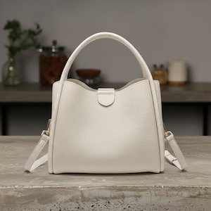 Könnte beinhalten: Eine cremefarbene Handtasche mit einem oberen Griff und einem abnehmbaren Schultergurt. Die Tasche hat ein strukturiertes Design mit gewölbtem Oberteil und kleinem Verschluss. Die Tasche ist aus strukturiertem Material.
