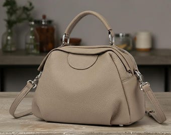 Puede incluir: Un bolso de mano color topo con forma redondeada, con asa superior y bandolera desmontable. El bolso tiene herrajes plateados y una superficie texturizada. El diseño incluye un cierre de cremallera y un pequeño detalle circular en la parte delantera.