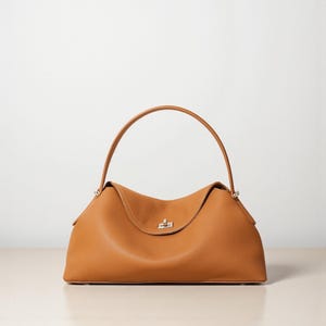 Puede incluir: Bolso de mano de cuero color marr&oacute;n claro con asa superior curvada y cuerpo estructurado. El bolso presenta un cierre de broche plateado y una sutil textura granulada. Dise&ntilde;o sencillo y elegante.