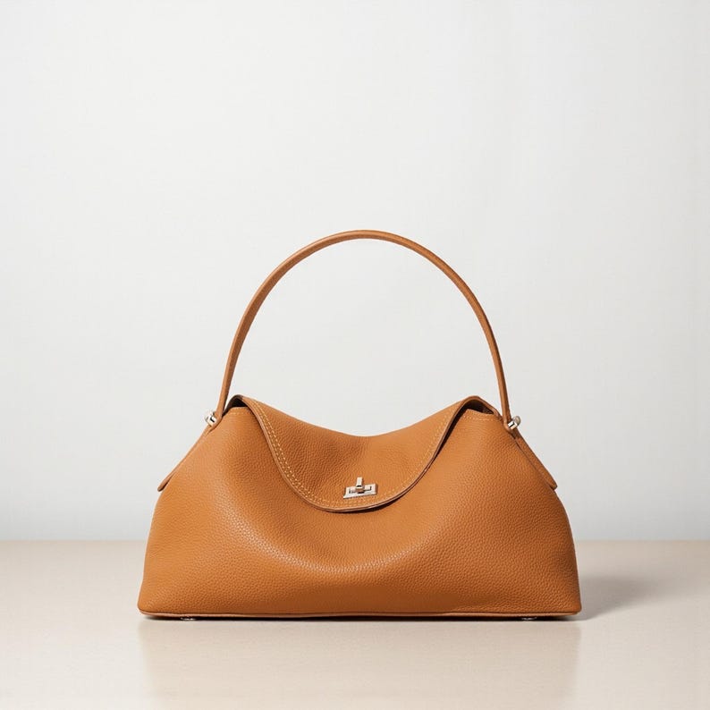 Puede incluir: Bolso de mano de cuero color marr&oacute;n claro con asa superior curvada y cuerpo estructurado. El bolso presenta un cierre de broche plateado y una sutil textura granulada. Dise&ntilde;o sencillo y elegante.