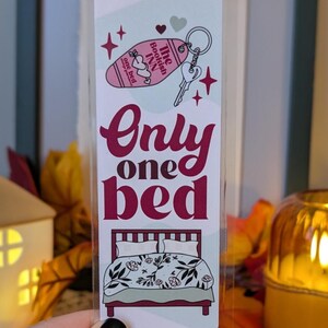 May include: A bookmark with the text "Only one bed" in burgundy script. The top features a pink hotel keychain with "The Bookish Inn" and a key. A bed with floral bedding is at the bottom. The bookmark is held in a hand.