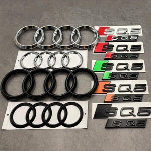 May include: An assortment of car emblems and badges, including chrome and black Audi rings, and SQ5 badges in various color schemes. The badges are on a light gray surface.