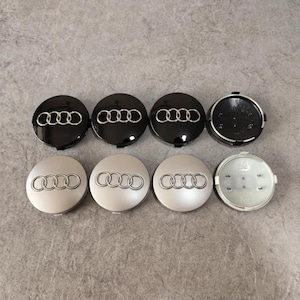 May include: Eight black and silver Audi wheel center caps. Four are black with a silver Audi logo, and four are silver with a silver Audi logo. One black cap is open, revealing the back.