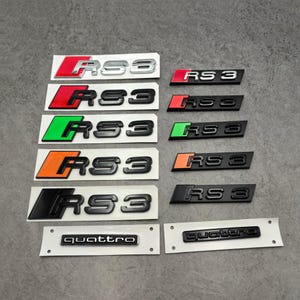 May include: A collection of RS3 and Quattro car emblems in various colors, including red, green, orange, and black. The emblems are on white backing and are displayed on a gray surface.