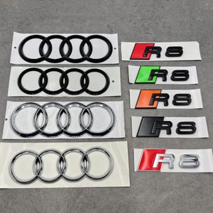 May include: A collection of Audi car emblems and R8 badges. The emblems feature the iconic four interlocking rings in black and chrome. The R8 badges come in various color schemes, including red, green, orange, black, and silver.