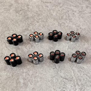 May include: A collection of black and silver tire valve stem caps. The caps feature the Audi logo in red and white. The caps are arranged in small groups on a gray surface.