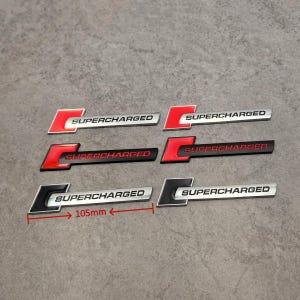 May include: A collection of six "SUPERCHARGED" emblem badges in various color schemes. The badges are approximately 4.1 inches long and feature a stylized "C" design. Colors include red, black, silver, and white. The badges are likely for automotive use.