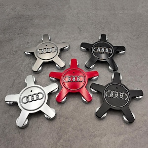 May include: Five car wheel center caps in silver, black, and red. Each cap features the Audi logo in the center and a five-point star design with silver accents. The caps are displayed on a gray surface.