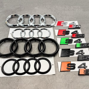 May include: An assortment of car emblems and badges, including chrome and black Audi rings, and S4 badges in various color schemes: red, green, black, and orange. The emblems are displayed on white backing.