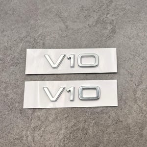 Puede incluir: Dos emblemas V10 plateados sobre un soporte blanco. Los emblemas, en fuente sans-serif, son probablemente accesorios automotrices. El fondo es una superficie gris texturizada.