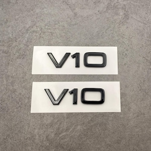 Puede incluir: Dos pegatinas negras con el emblema V10 sobre un soporte blanco. Los emblemas son brillantes y tienen una apariencia tridimensional. El fondo es una superficie gris neutra.