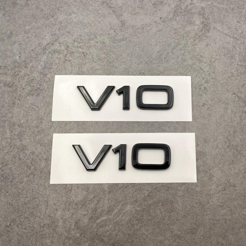 Puede incluir: Dos pegatinas negras con el emblema V10 sobre un soporte blanco. Los emblemas son brillantes y tienen una apariencia tridimensional. El fondo es una superficie gris neutra.
