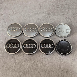 May include: Eight silver and black Audi wheel center caps. The caps have the Audi logo in the center, with a silver ring around the edge. Some caps are silver, and some are black.
