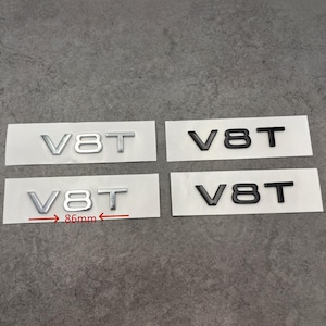 May include: Four V8T emblem badges, two in silver and two in black, each mounted on a white backing. The silver badges measure 86mm in width. The emblems are designed for automotive use.