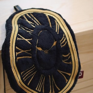 May include: A navy blue, plush clock-shaped item with gold Roman numerals and a gold border. The clock face features gold hands and a central circle. A black strap is attached to the top.