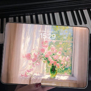Könnte beinhalten: Ein Tablet zeigt einen floralen Hintergrund mit rosa Rosen in einer grünen Vase an einem Fenster. Die Uhrzeit 15:29 und das Datum werden angezeigt. Das Tablet wird vor einer Klaviertastatur gehalten.