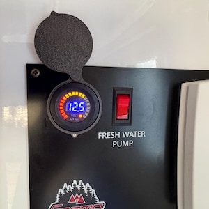 Puede incluir: Un panel negro con un voltímetro digital que muestra 12,5 voltios, un interruptor rojo con la etiqueta "FRESH WATER PUMP" y un logotipo. El voltímetro tiene una cubierta protectora. Un termostato blanco es visible a la derecha.