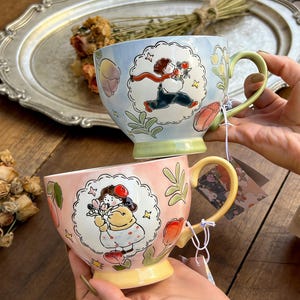 May include: Two ceramic mugs with whimsical illustrations. One mug is light blue with a running figure carrying flowers, the other is pink with a girl holding flowers. Both mugs have colorful floral and leaf designs, with green and yellow handles.