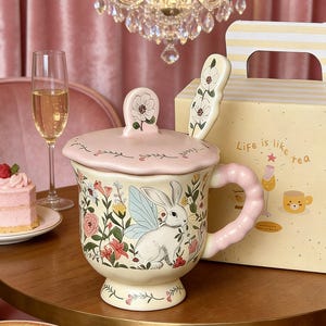 Puede incluir: Una taza de té de cerámica con tapa, cuchara y caja de regalo. La taza es de color crema con un diseño floral y de conejito. La tapa y el asa son rosas. La cuchara tiene un diseño floral. La caja de regalo tiene el texto "Life is like tea".
